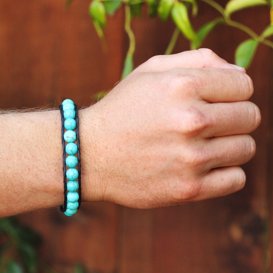 Close-up of a man's wrist wearing a cracked turquoise wrap bracelet with a blurred brown wood with green tree leaves in the background.