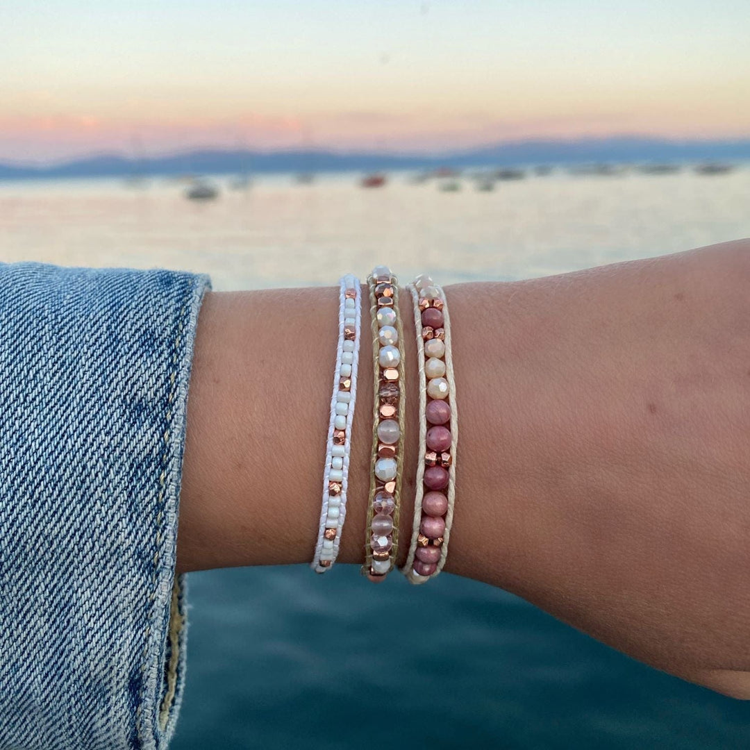 Close-up of a woman's wrist with a blurred lake in the background, wearing a blue denim jacket, and three wrap style bead bracelets, in pink, white, rose gold, and cream colors.