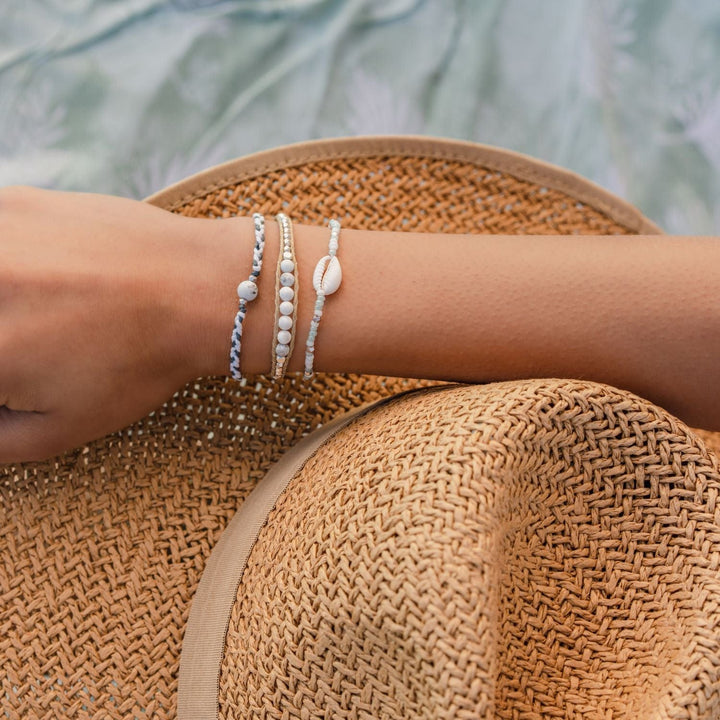 Close-up of the back of a woman's head wearing a straw sun hat, with her arm across the top of the hat, wearing three bracelets on her wrist, in shades of white, gray, tan, and blue, one with a single white sea shell.