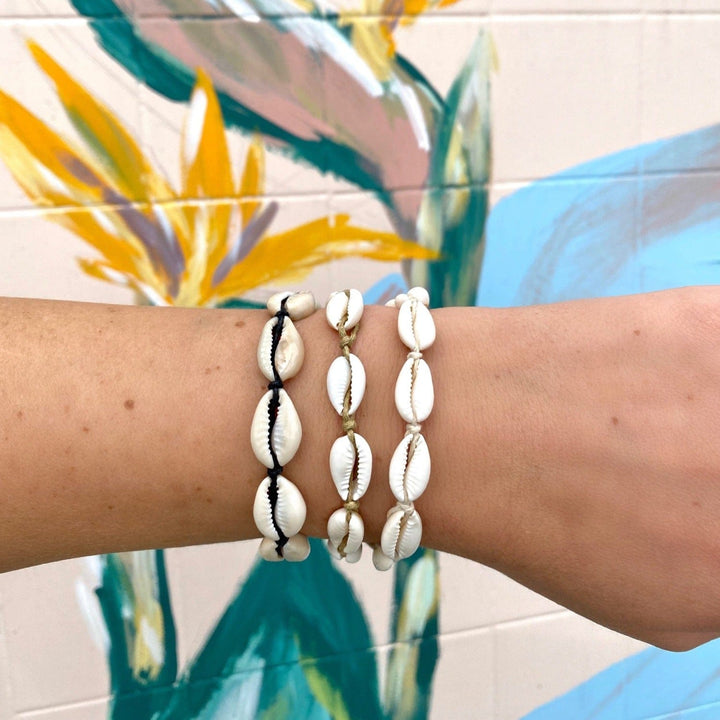 Close-up of a woman's wrist with three cowry shell bracelets, one with black cord, one with white cord, one with tan cord, with a wall painted with flowers in pastel colors.