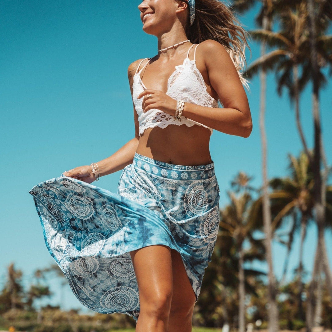 Young woman with long blonde hair, wearing a white lace halter crop top and a blue and white print skirt blowing in the wind as she's running on the beach, wearing cowry shell bracelets on her wrist, with blurry green trees and blue sky in the background.