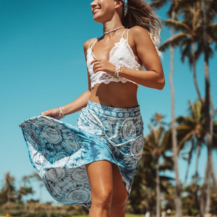 Young woman with long blonde hair, wearing a white lace halter crop top and a blue and white print skirt blowing in the wind as she's running on the beach, wearing cowry shell bracelets on her wrist, with blurry green trees and blue sky in the background.