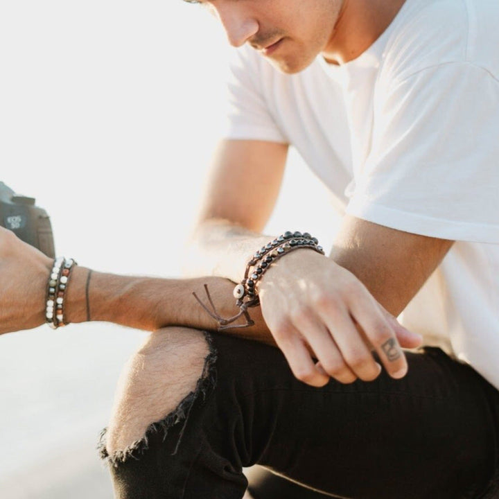 Close up of a young man's wrists wearing two wrap style bead bracelets on each wrist, in shades of brown, black, and white, including the wipeout hematite bracelet.