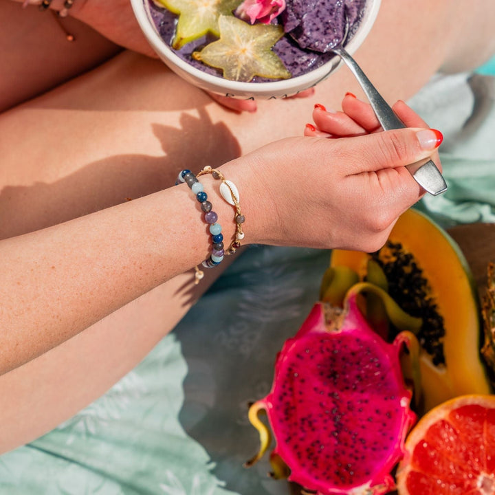 Close-up of a woman's legs and hand, holding a spoon in a bowl of purple blueberry yougurt with yellow star fruit on top, sitting outside on a beach towel with bare legs, wearing two bead bracelets, one a sea shell bracelet, and the wisdom vitality healing bracelet, with a side display of half a pink dragon fruit, papaya, and red orange in a wood bowl.