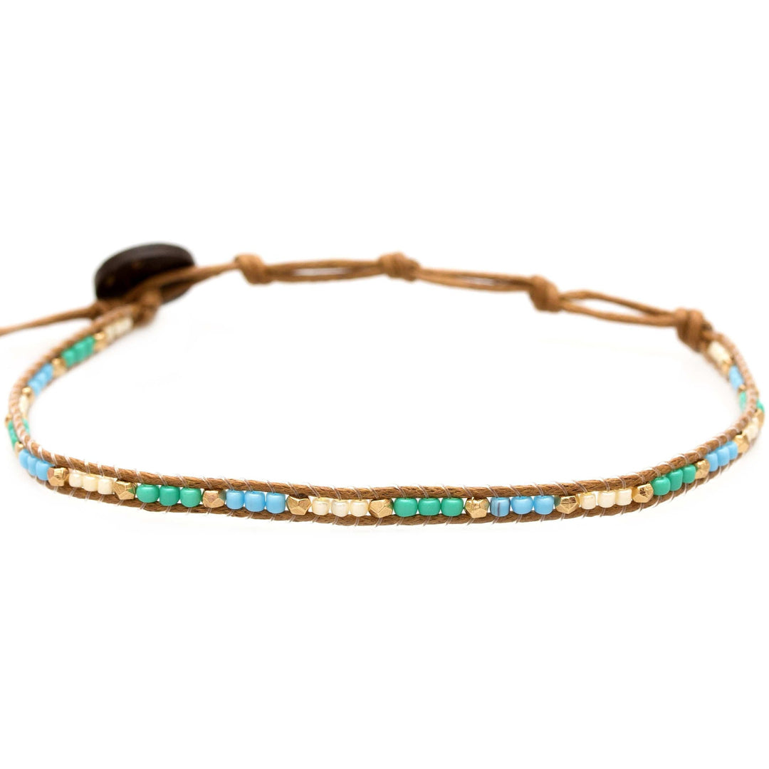 Close-up of a seed bead ankle bracelet made with pearl white, turquoise, and gold seed beads, with an adustable brown colored cord and a dark brown coconut shell button closure.