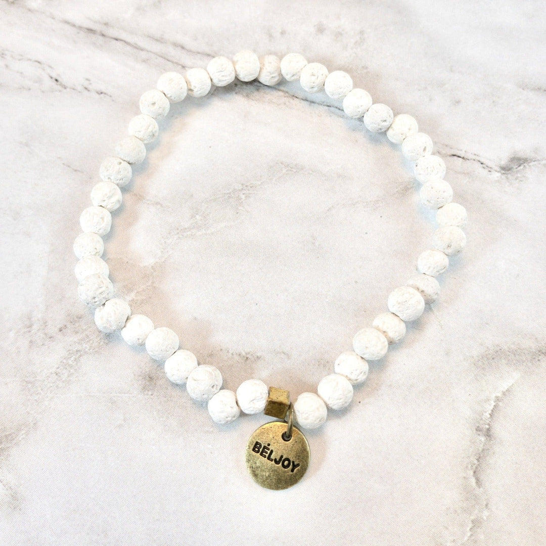 A stretch style white lava stone bead bracelet with 4mm round beads, a square gold brass bead accent, and a gold brass beljoy logo charm, displayed on a white and gray marble background.