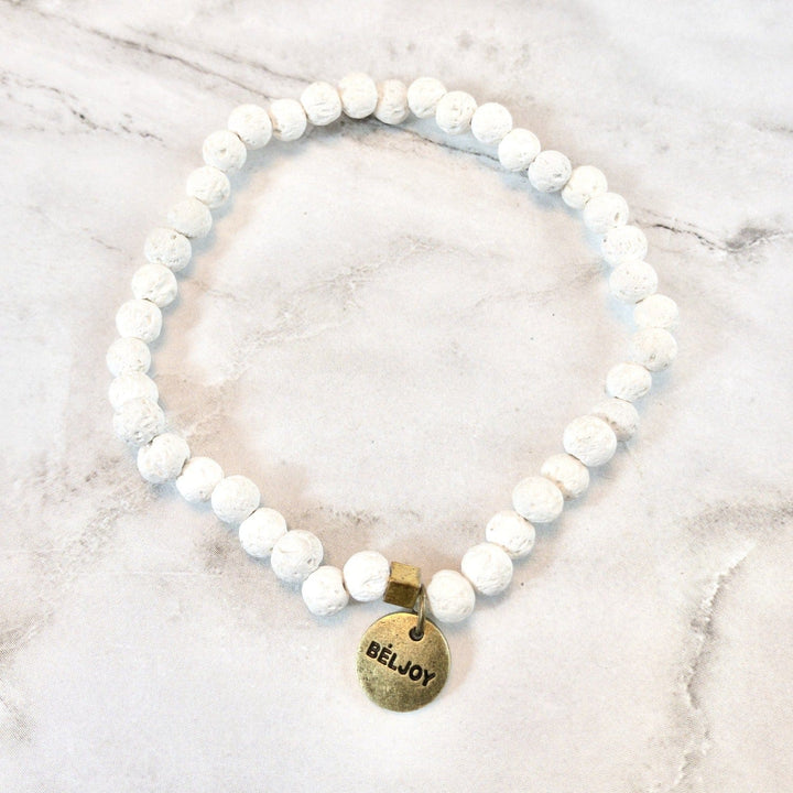 A stretch style white lava stone bead bracelet with 4mm round beads, a square gold brass bead accent, and a gold brass beljoy logo charm, displayed on a white and gray marble background.